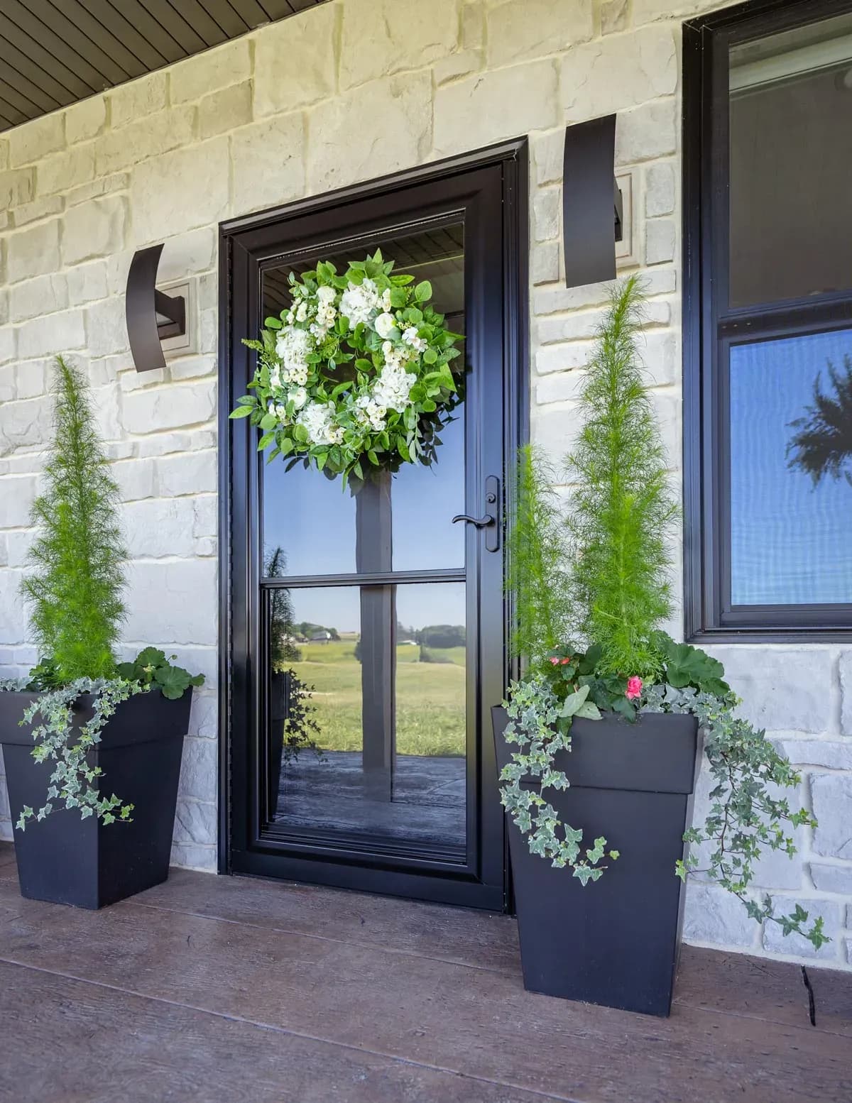 A black-framed glass door adorned with a lush green wreath featuring white flowers is flanked by two tall planters with greenery, set against a stone wall.