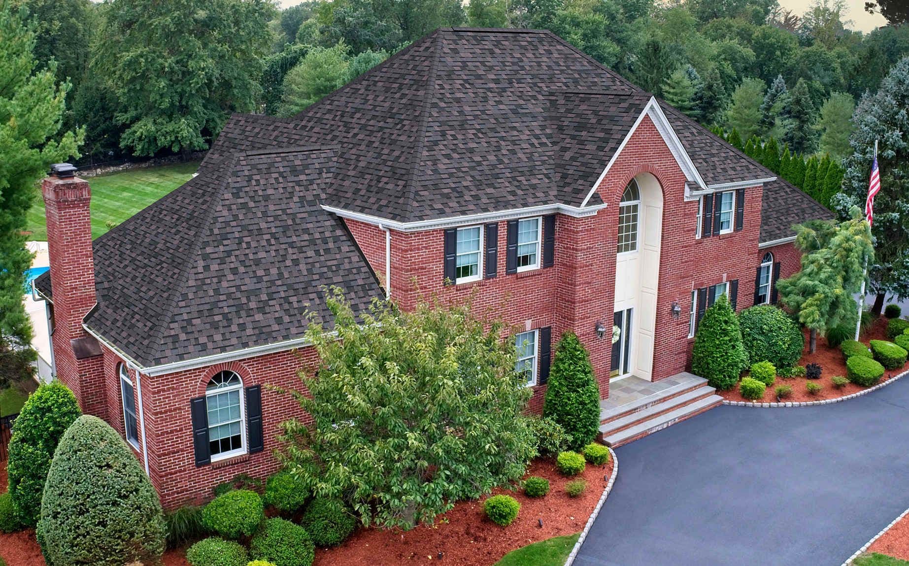 Aerial view of a two-story brick colonial home with dark shingle roof, curved driveway, brick chimney, and manicured landscaping surrounded by trees