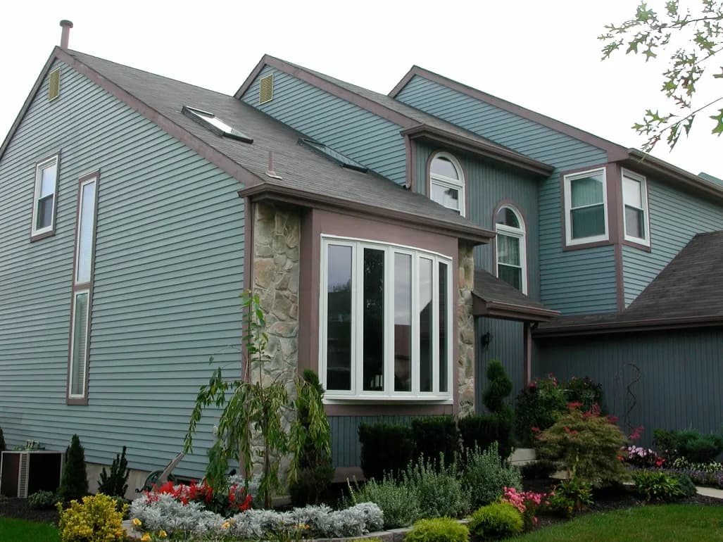 A charming, modern house with light blue siding, stone accents, large windows, and a well-kept garden. The scene conveys a peaceful, inviting atmosphere.