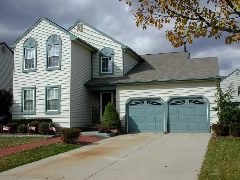 Two-story house with light siding and teal trim, featuring a double garage and a tidy lawn. The sky is overcast, creating a calm suburban feel.