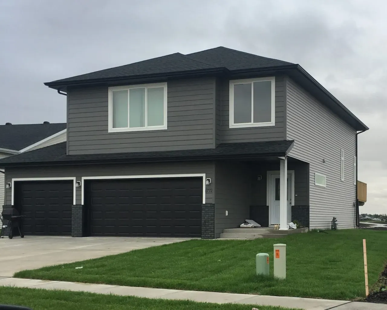 Modern two-story gray house with a dark roof, three black garage doors, and a well-maintained green lawn. The sky is overcast, creating a calm atmosphere.