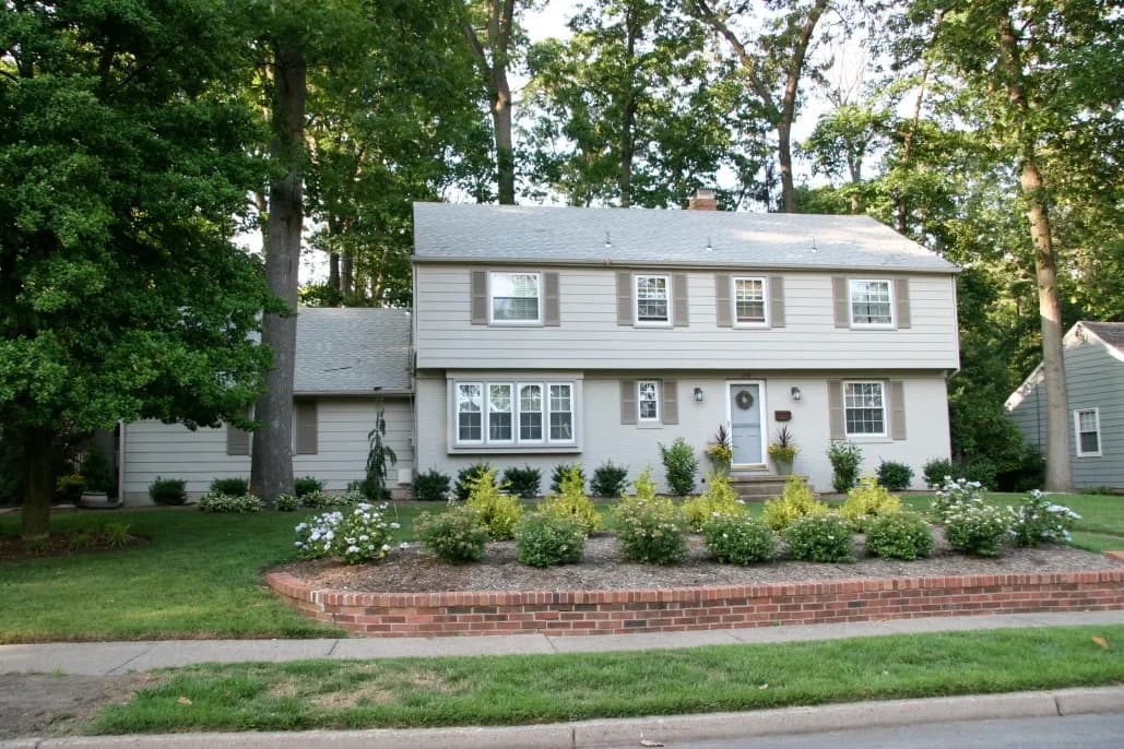 Two-story suburban house with a gray exterior, white trim, surrounded by lush greenery and trees. The garden features flower beds and a brick border.