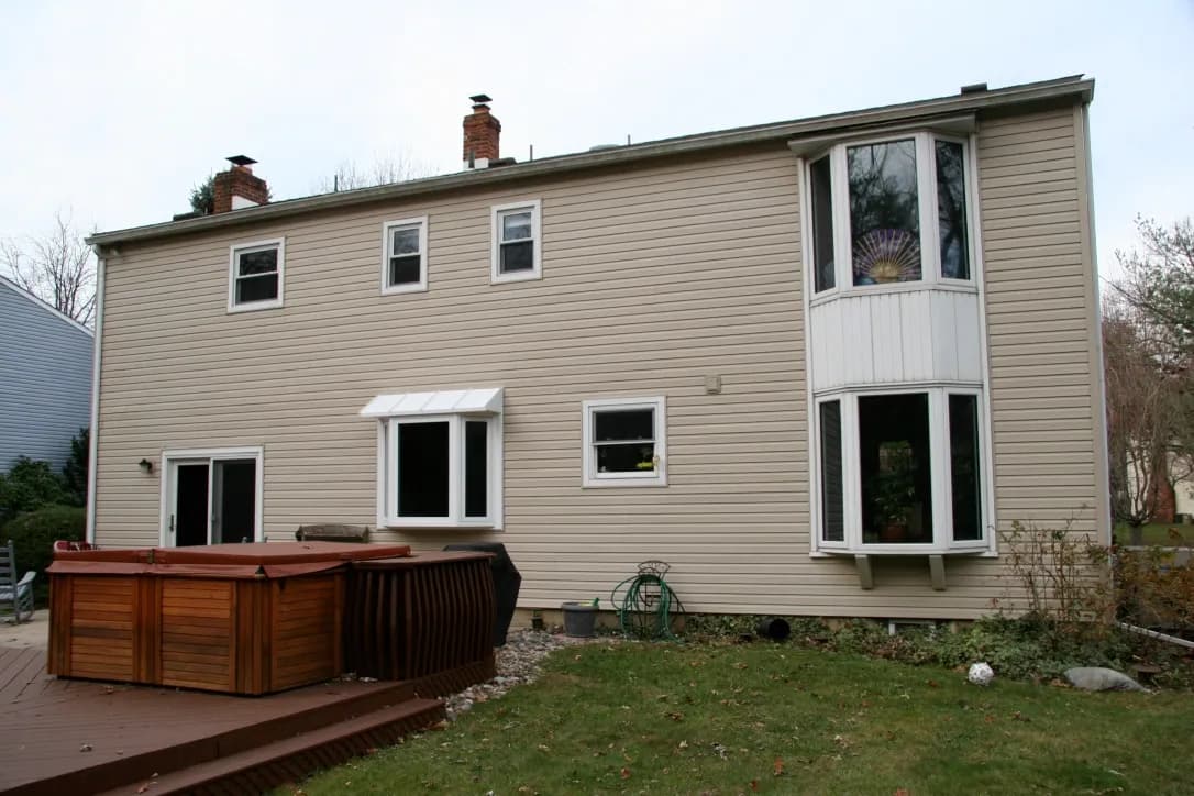 Two-story beige house with a slanted roof, four small windows, and a large bay window. In the backyard, a closed wooden hot tub sits on the deck, with grass and bushes nearby. The scene is calm, suggesting a suburban setting.