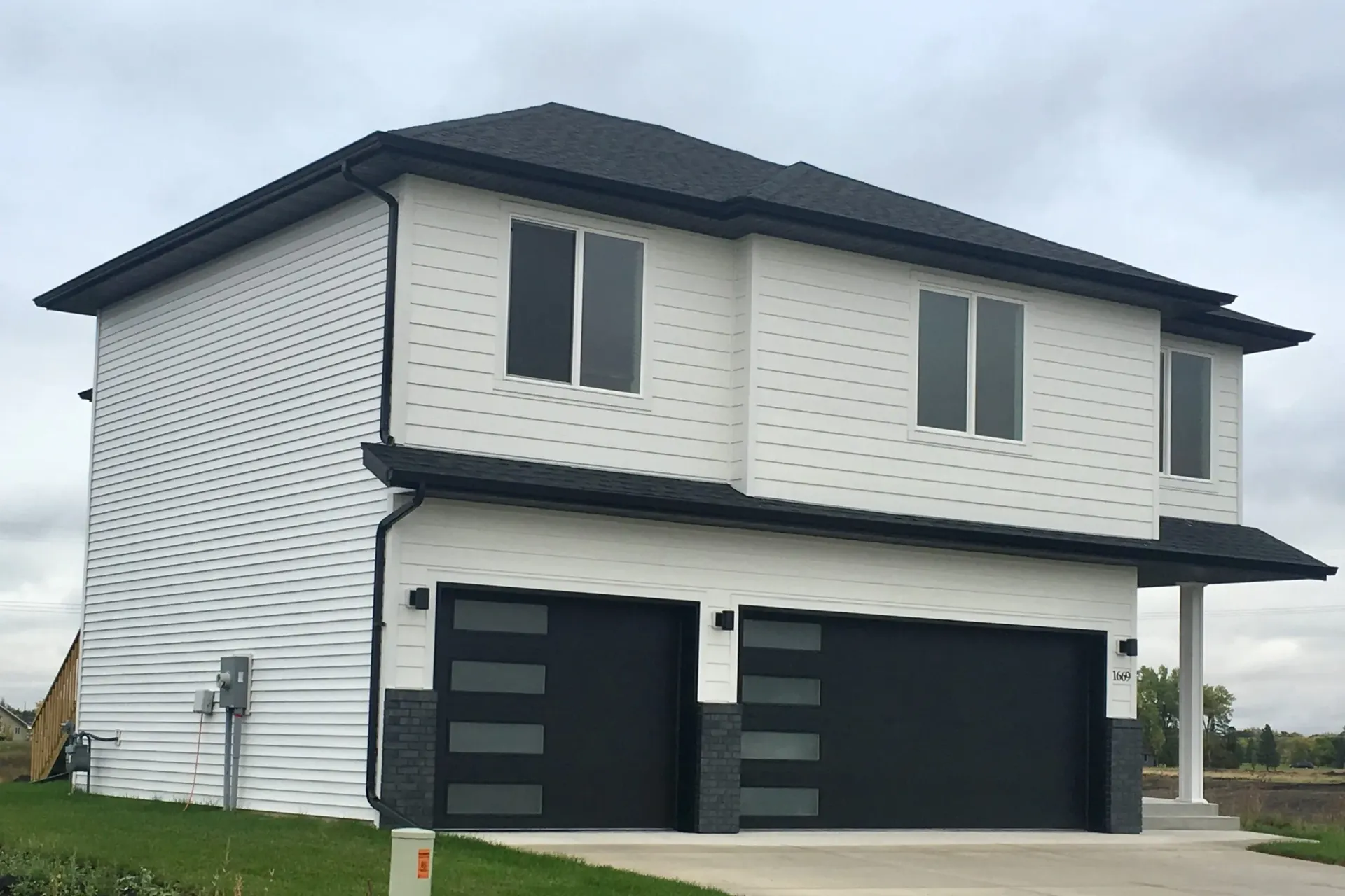 Modern two-story house with white siding and black roof. Features include large garage doors, rectangular windows, and a clean, minimalist design.