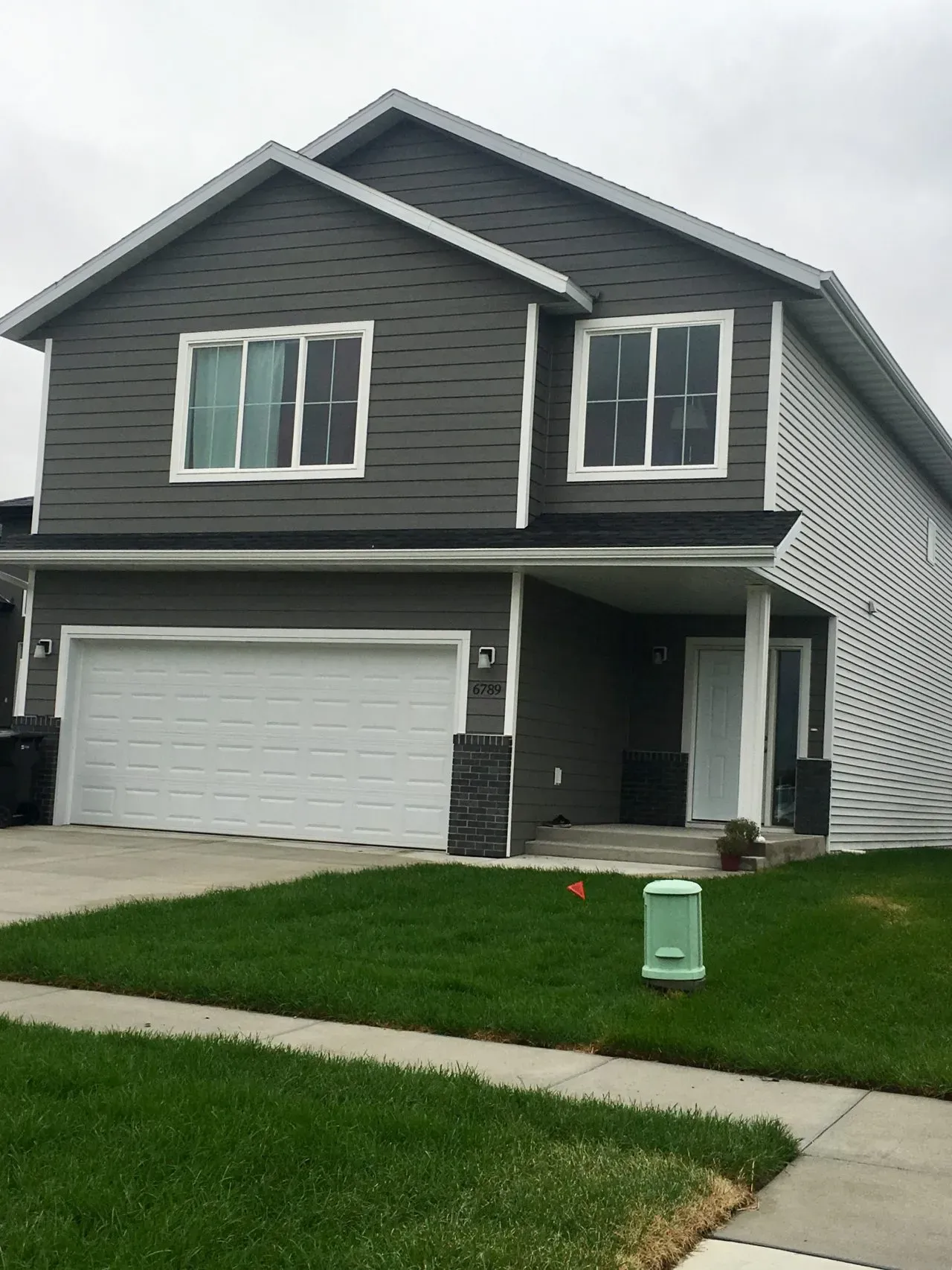 Two-story suburban house with gray siding, white trim, and a double garage. Neatly trimmed green lawn, a small porch, and a cloudy sky overhead.