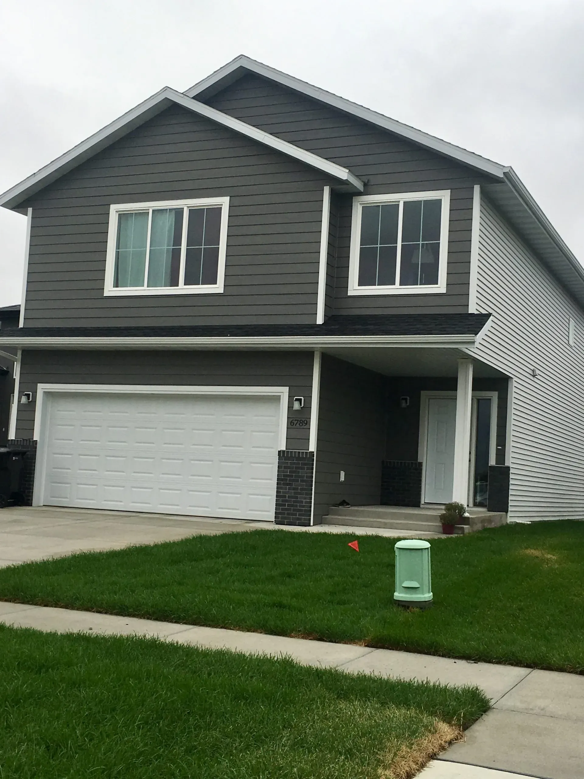 Two-story suburban home with gray siding and white trim, featuring a double garage and a green, well-kept lawn. The atmosphere is calm and residential.