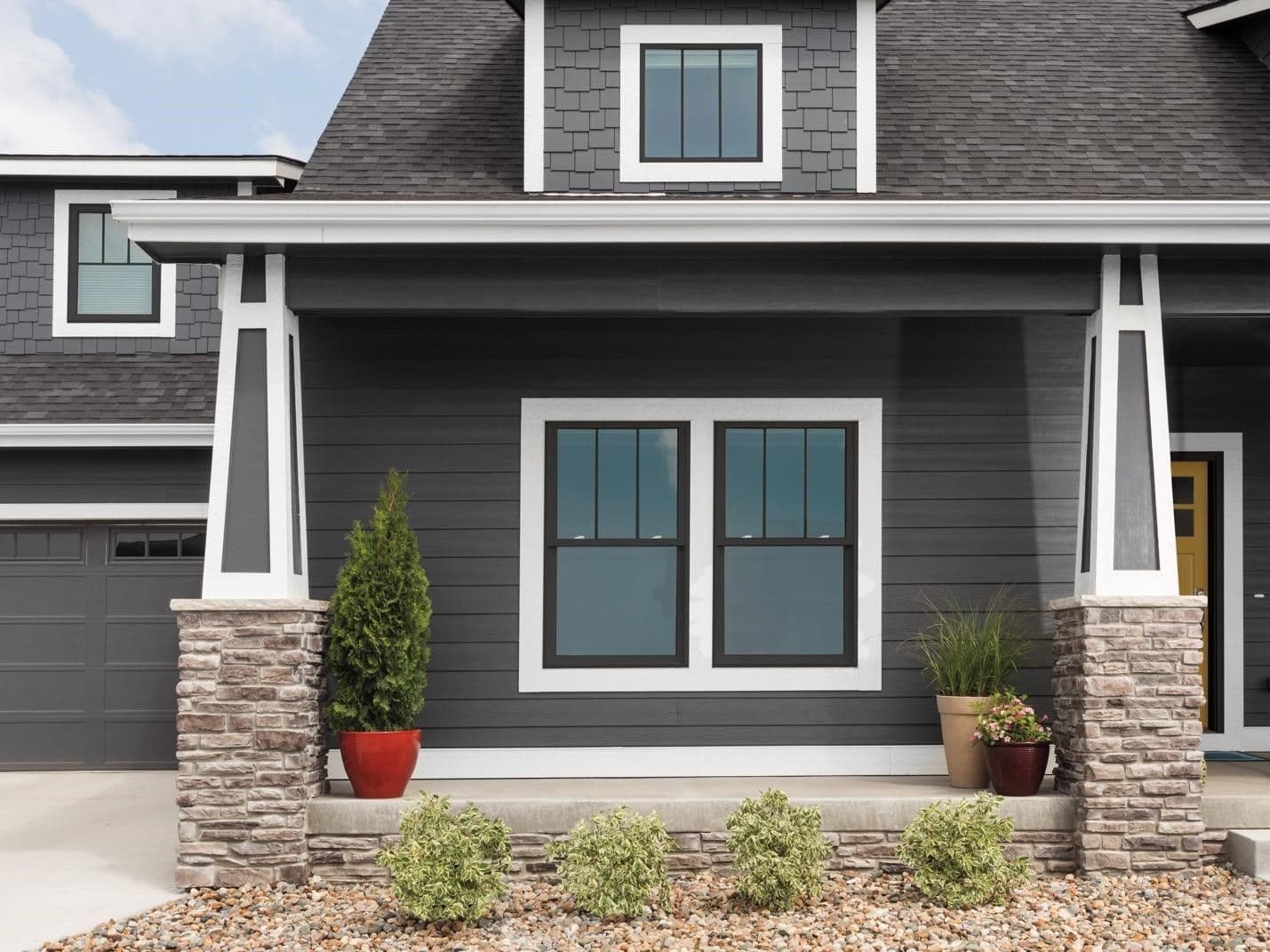 Modern house exterior featuring dark gray siding, white window trim, and pillars with stone bases. A porch with potted plants adds a welcoming touch.