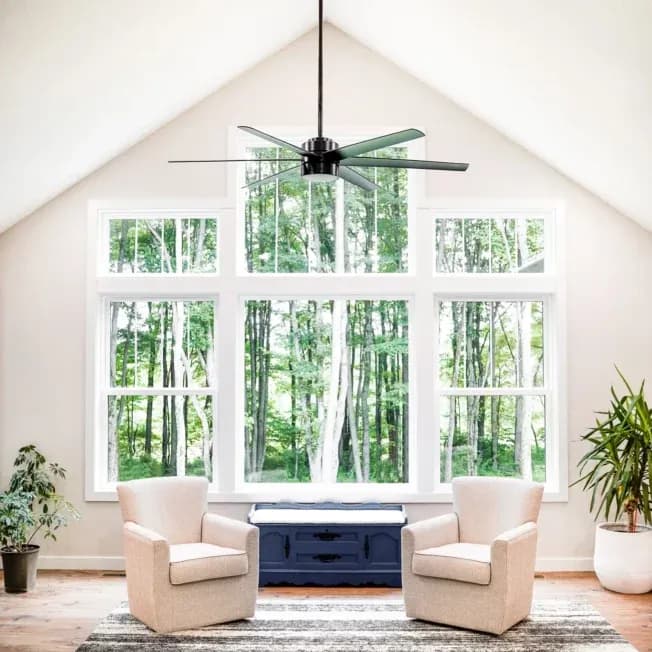 Bright living room with a high ceiling, large windows showing a lush forest. Two beige armchairs face each other, a blue cabinet between them.