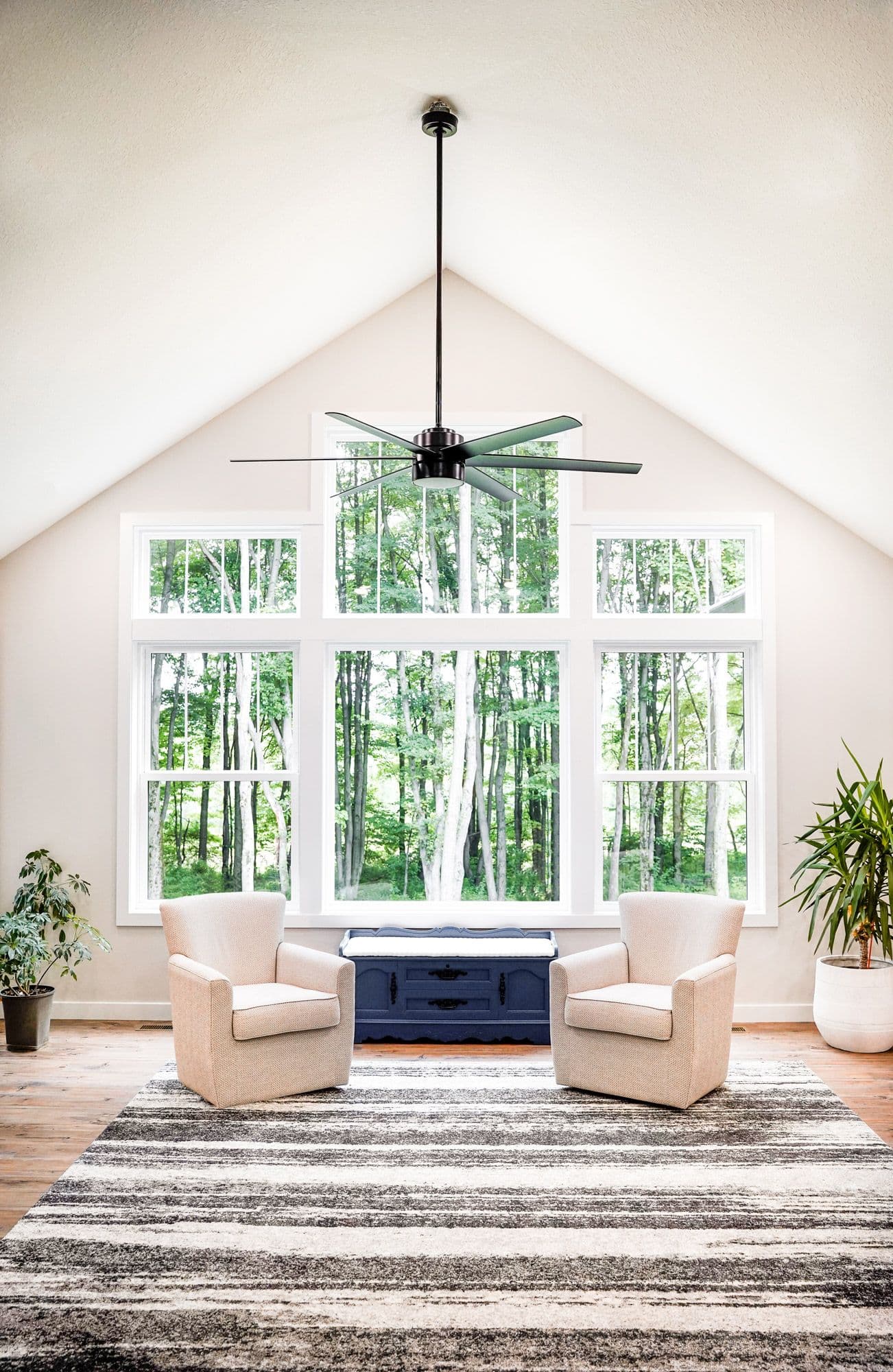 Bright room with large windows overlooking a forest, two beige armchairs, a striped rug, blue chest, plants, and a black ceiling fan. Peaceful and airy.
