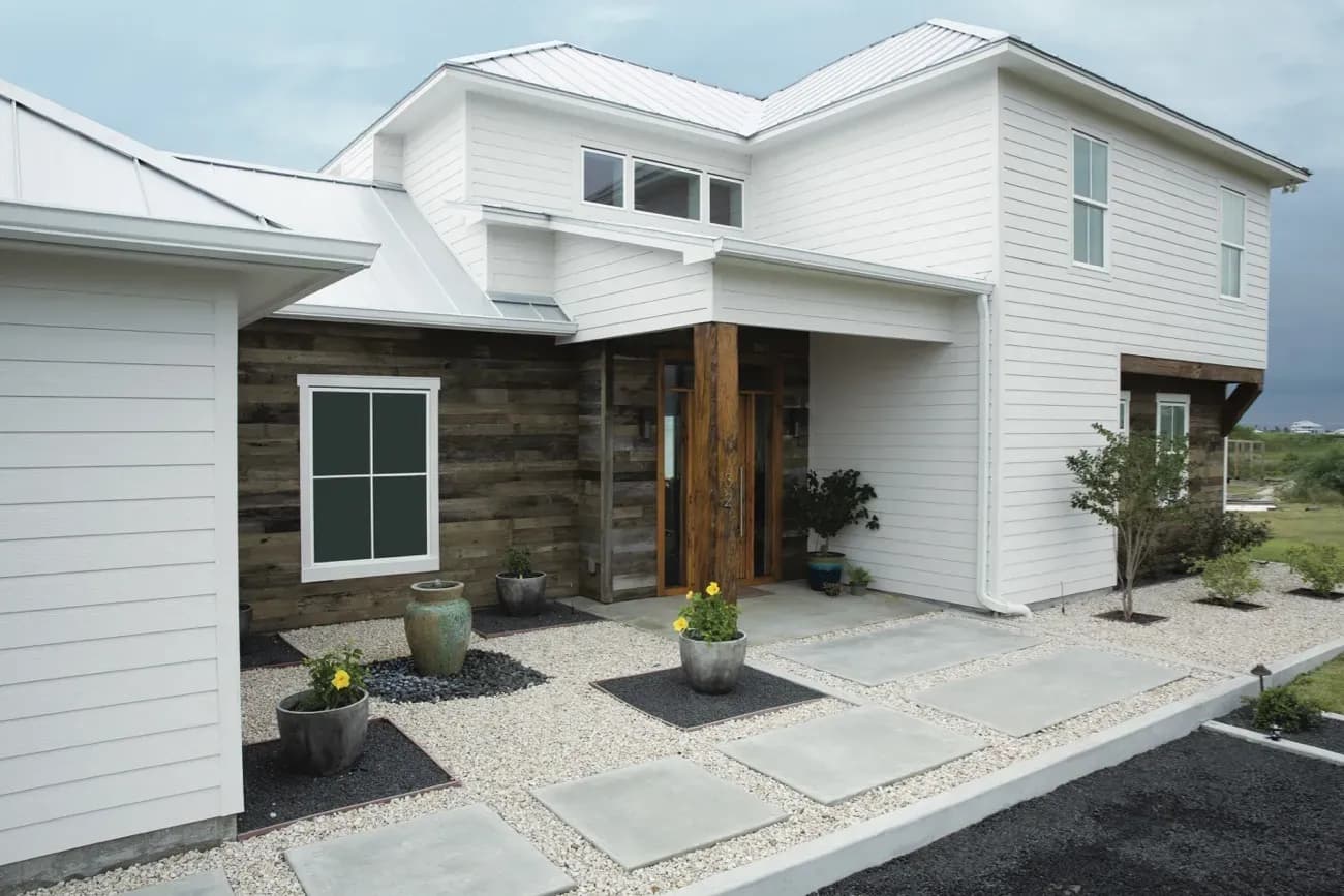 Modern white house with wood accents, a metal roof, and large windows. The front yard features potted plants and a stone pathway, set against a cloudy sky.