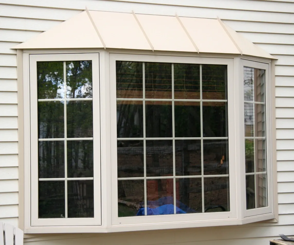 A large bay window with white frames and gridded panes on a white siding house. The window reflects greenery, suggesting an outdoor view.