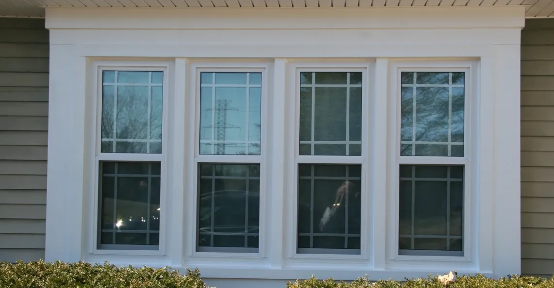 A set of three large white-framed windows on a beige house exterior. Reflections and trees are visible through the glass, with bushes below the windows.