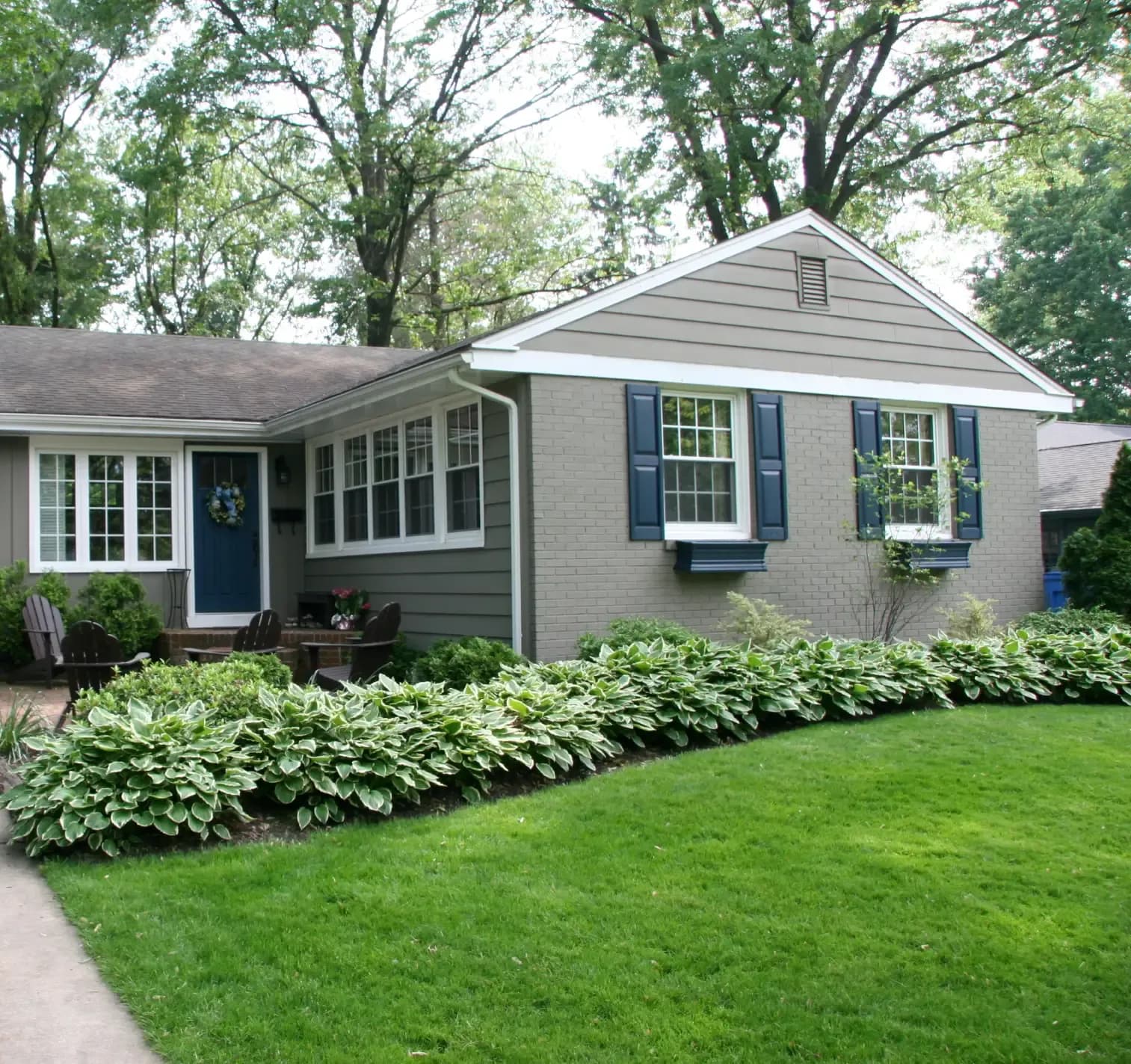 Charming, gray single-story house with blue shutters, lush green lawn, and a hosta-lined pathway. Large trees provide a serene backdrop.