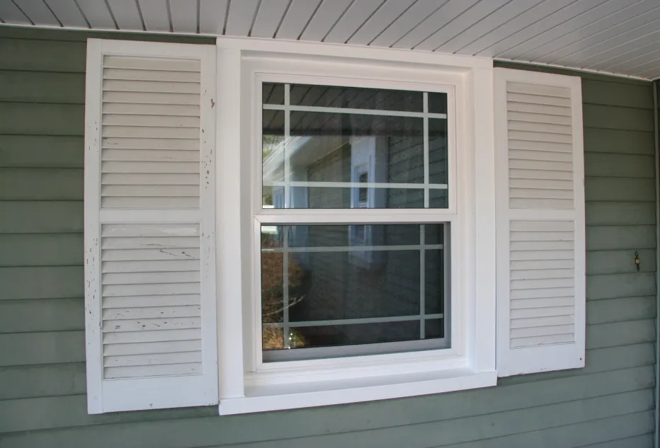 White-framed window with shutters, set in light green siding. The window's reflection shows a sunny yard, conveying a calm and homey atmosphere.