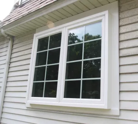 A white-framed, double-pane window with grid design is set in beige siding, reflecting greenery and a partly cloudy sky, conveying a serene ambiance.