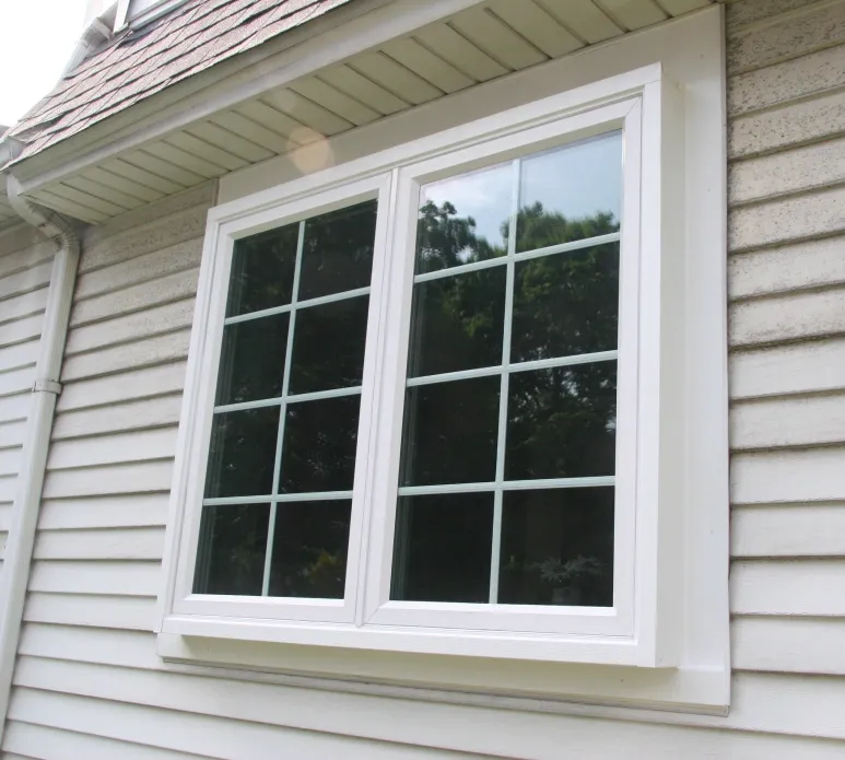 White-framed double window with grid design on a light gray siding house, beneath an overhang. The glass reflects greenery from a nearby tree.