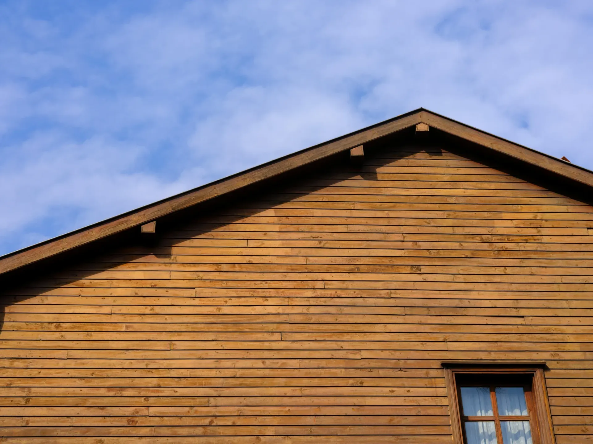 Gable end of a wooden house with horizontal planks, a single window, and a clear blue sky above, conveying a serene and rustic atmosphere.