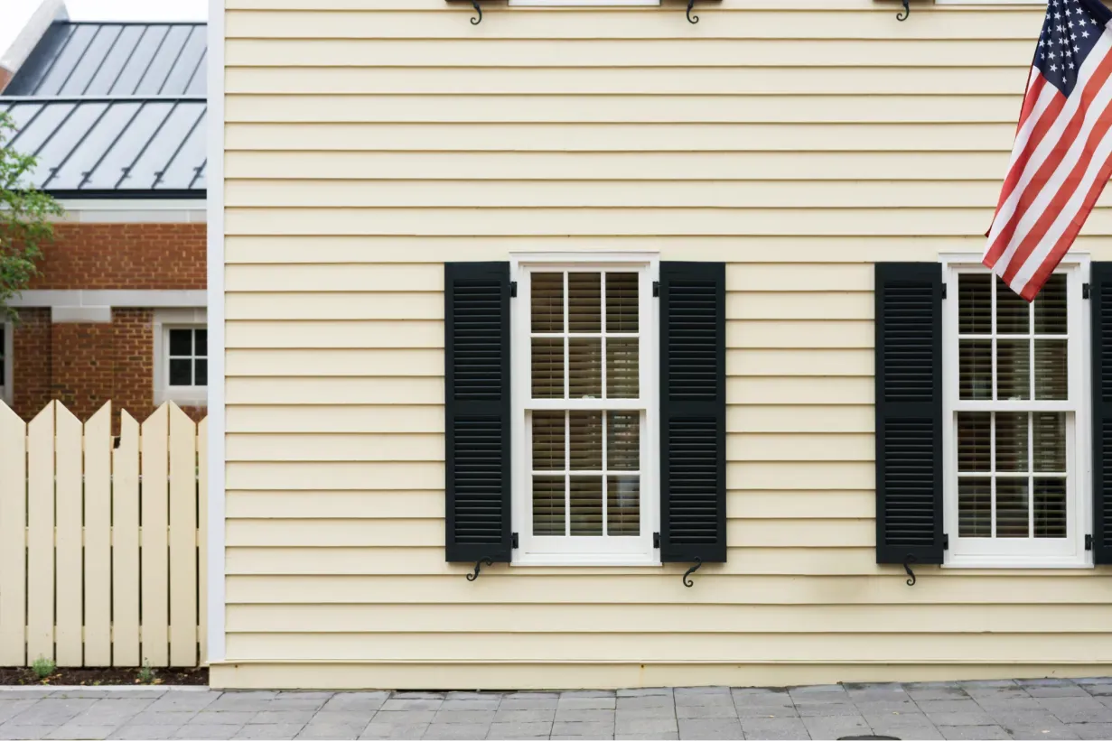 Side of a beige house with black shuttered windows, an American flag, and a white picket fence. The scene conveys a classic, suburban feel.