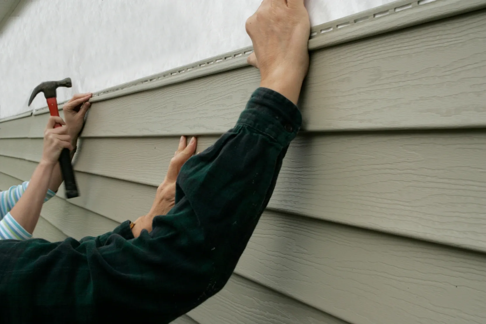 Two people are installing gray vinyl siding on a house. One person aligns the siding, while the other secures it in place with a hammer.