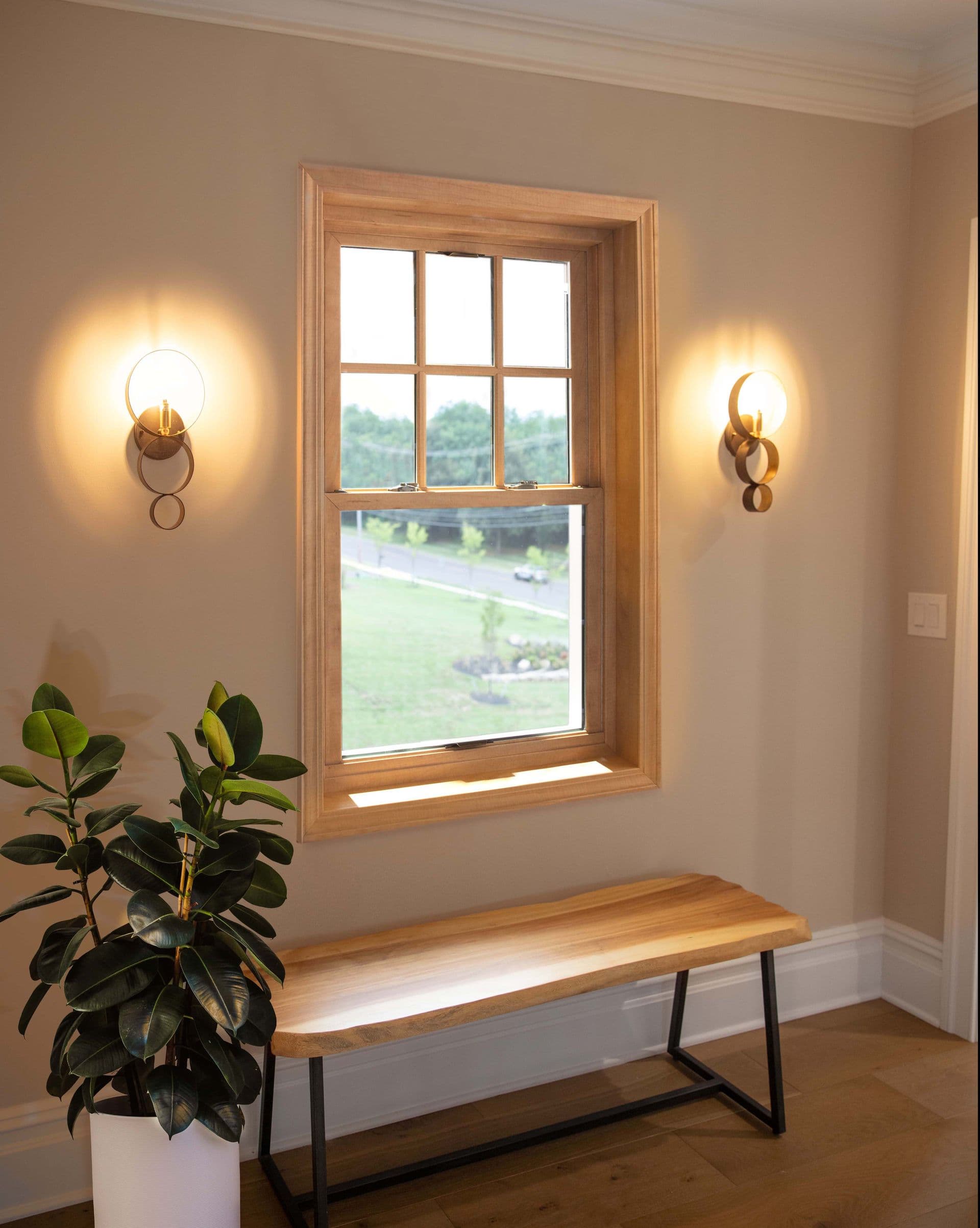 A serene room with a wooden window frame showing a view of a green landscape. Two modern wall sconces emit warm light. A sleek wooden bench sits below, and a lush potted plant beside it adds a touch of nature.