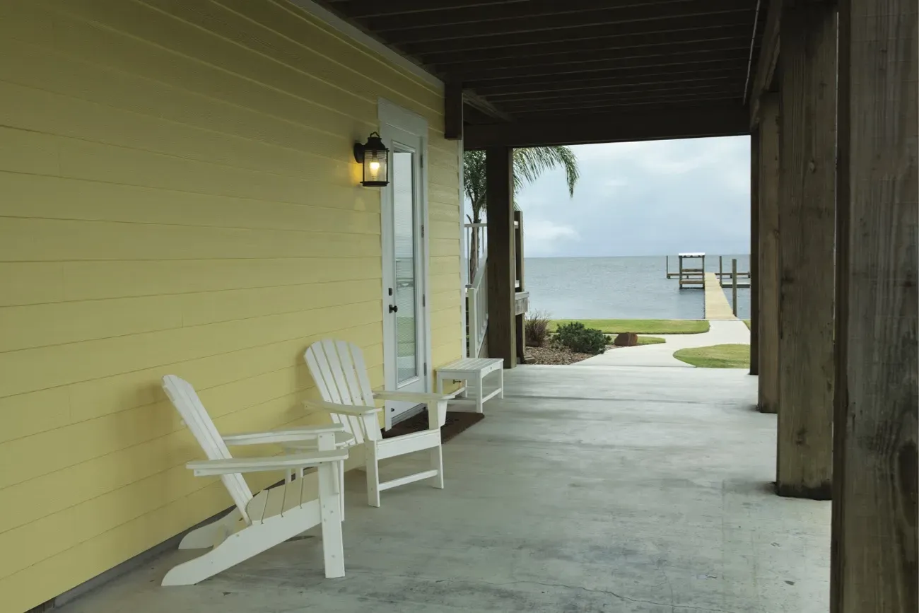 Covered patio with yellow siding features two white Adirondack chairs. It leads to a wooden pier extending over a calm lake, creating a serene atmosphere.